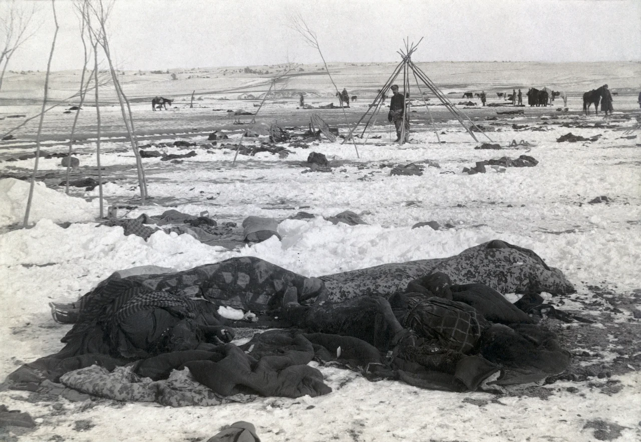 Bibliographic caption: "Big Foot's camp three weeks after Wounded Knee Massacre; with bodies of four Lakota Sioux wrapped in blankets in the foreground; U.S. soldiers amid scattered debris of camp" 1 photographic print : albumen ; 9 x 11 in.